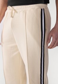 Cremefarvede sweatpants med snøre i taljen og to lodrette sorte og hvide striber langs siderne. Blød tekstur, afslappet pasform.