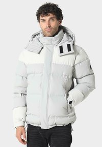 Vaaleanharmaa toppatakki valkoisilla yksityiskohdilla. Siinä on korkea kaulus, säädettävä huppu, sivutaskut ja joustavat hihansuut. Teksturoidusta kankaasta.