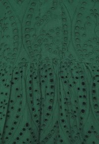 Grüner Stoff mit kunstvoll gestickten Mustern und ausgeschnittenen Löchern, die ein strukturiertes und detailliertes Erscheinungsbild erzeugen.