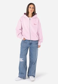 Rózsaszín cipzáras kapucnis pulóver nagy kapucnival és elülső zsebekkel, amit laza szabású kék farmernadrággal párosítanak, amelyen egy szakadt folt található, valamint fehér sportcipővel.