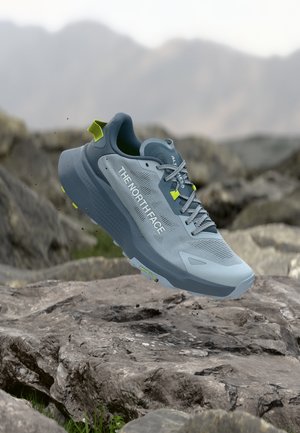 Zapatilla de trail running The North Face en azul claro y gris con acentos amarillos, suspendida sobre terreno rocoso al aire libre.