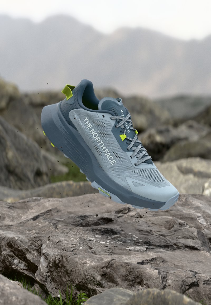 Ανοιχτό μπλε και γκρι παπούτσι για τρέξιμο σε μονοπάτι The North Face με κίτρινες λεπτομέρειες να αιωρείται πάνω από βραχώδες έδαφος σε εξωτερικό χώρο.