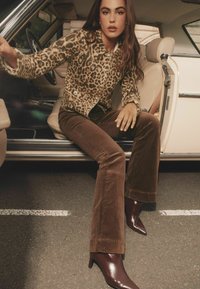 Wolljacke mit Leopardenmuster und goldenen Knöpfen, braune Corduroy ausgestellt-Hosen und glänzende burgunderfarbene Stiefeletten mit spitzen Zehen, sitzend in einem Auto.
