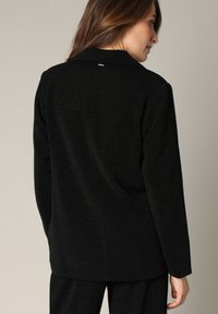 Zwarte blazer met een subtiele glans, voorzien van een klassieke kraag, lange mouwen en een strakke, op maat gemaakte pasvorm. De achterkant heeft een klein splitdetail.