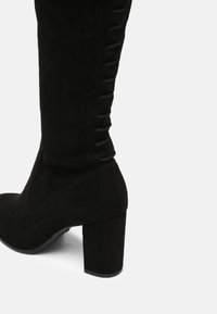 Černý vysoký kožený bot s hladkým povrchem, s vertikálním šněrováním na bocích a širokým podpatkem.