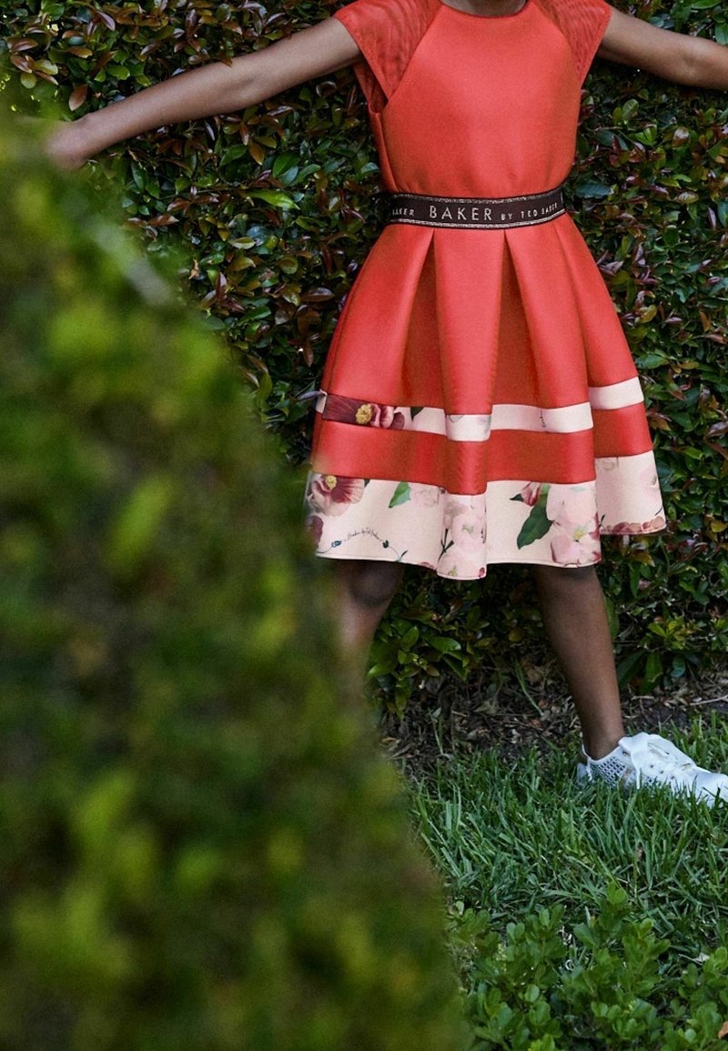 Enfant en robe rouge avec bordure florale et baskets blanches, debout les bras écartés contre une haie verte feuillue.