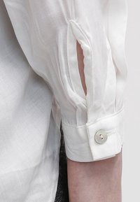 Bílá košile s rukávy na knoflík, která má jedinečný rozparek. Látka je průhledná a hladká s mírným leskem.