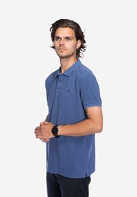 Blauwe poloshirt van zacht katoen, met een drieknopenlijst en geborduurd logo op de linkerborst, gedragen met donkere broek.