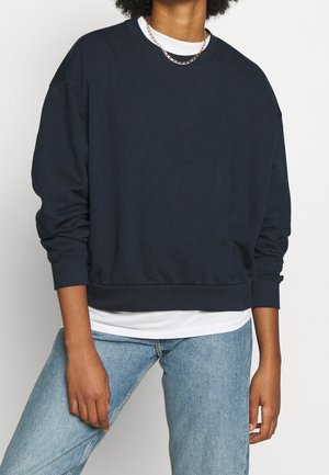 Personne portant un sweat-shirt oversize bleu marine foncé par-dessus une chemise blanche, associé à un jean bleu clair et un collier chaîne en or.
