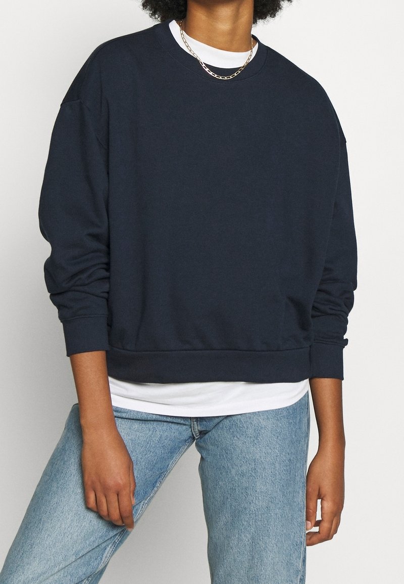 Personne portant un sweat-shirt oversize bleu marine foncé par-dessus une chemise blanche, associé à un jean bleu clair et un collier chaîne en or.