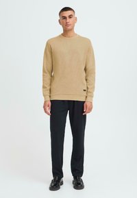 Maglietta a manica lunga in maglia waffle beige con polsini e orlo a costine, abbinata a pantaloni neri a gamba stretta e scarpe nere. Design semplice e moderno.