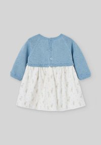 Cardigan bleu en tricot avec des manches longues, boutonné à l'arrière, associé à une robe blanche ornée d'un motif floral de petits vases.