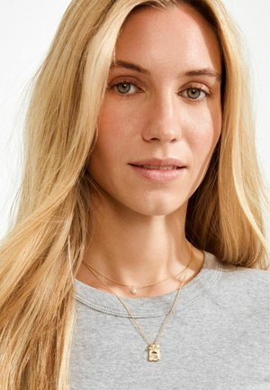 Kihilised kullast kaelakeed ripatsi ja väikese pärliga, kantud üle halli pikaahelalise kampsuni. Juuksed on sirged ja blondid, nägu raamistavad.