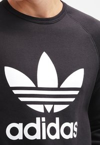 Svart sweatshirt med en stor vit Adidas Trefoil-logotyp med tre ränder undertill, rund halsringning och textilstruktur.