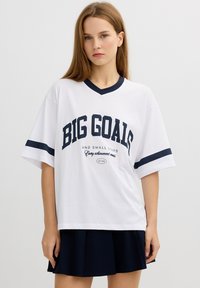 Camiseta blanca de gran tamaño con acentos en azul marino, que presenta "GRANDES METAS" en letras grandes. Mangas cortas y diseño de cuello en V.