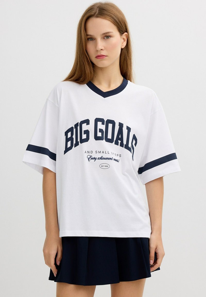 Camiseta blanca de gran tamaño con acentos en azul marino, que presenta "GRANDES METAS" en letras grandes. Mangas cortas y diseño de cuello en V.