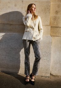 Ekranowy żakiet z mieszanki wełny z piórowymi akcentami przy mankietach, zestawiony ze srebrnymi wzorzystymi spodniami i czarnymi szpilkami z paskami wokół kostki.