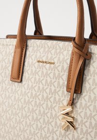 Borsa a mano in tessuto beige con dettagli in pelle marrone, caratterizzata da un motivo geometrico e un charm dorato "MK". Logo dorato "MICHAEL KORS" in evidenza.