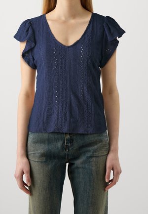 Femme portant un chemisier bleu marine texturé à manches courtes avec des manches volantées et un jean bleu délavé sur un fond uni.