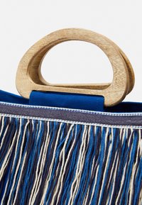 Blå och marinblå tygväska med ett runt handtag i trä, med långa fransar i blått, marinblått och krämfärgat. Sytt vita detaljer synliga.