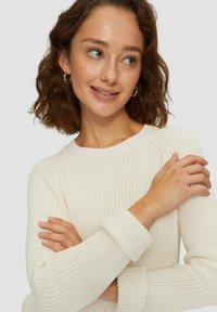Pull en tricot crème, avec un col rond, des poignets côtelés et un tissu texturé. Le modèle a les bras croisés, mettant en valeur les détails du pull.