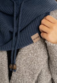 Marineblauwe gebreide sjaal met een geribde textuur, verstelbare trekkoorden met houten kralen, en een tan kleurige label met het merk "KNIT FACTORY".