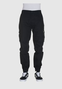Zwarte cargobroek met elastische pijpen, zijzakken en een knoopsluiting. Gedirigeerd met zwarte sneakers met witte accenten.