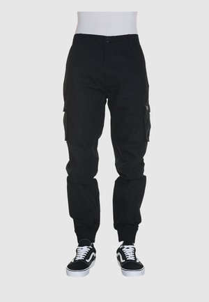 Pantaloni cargo neri con polsini elasticizzati, tasche laterali e chiusura a bottone. Indossati con sneakers nere con dettagli bianchi.