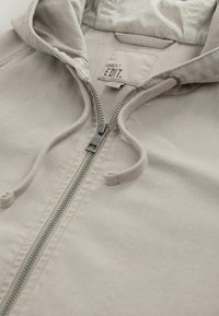 Sweat-shirt à capuche beige clair avec une fermeture éclair métallique à l'avant, des cordons de serrage, et une étiquette visible "NEXT EDIT." à l'intérieur du col.