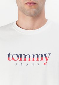 Hvit bomulls T-skjorte med teksten "tommy" i fet rød og marineblå, med "JEANS" under i en mindre marineblå skrift. Enkel rund hals.