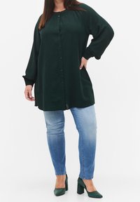Blusa lunga a maniche con bottoni sul davanti, di colore verde scuro, con colletto arricciato e polsini elasticizzati. Abbinata a jeans skinny azzurri e scarpe con tacco a blocco verdi.