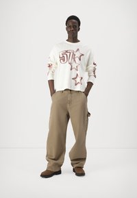 Maglione bianco a maglia con motivo a stelle color bordeaux e una grande scritta '57', abbinato a pantaloni beige larghi e stivali alla caviglia marroni.