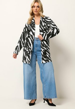 Vrouw draagt een zwart-wit zebraprint blazer, witte top, lichtblauwe jeans met wijde pijpen en zwarte hakken, staand tegen een effen achtergrond.