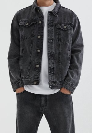 Mężczyzna ubrany w czarną kurtkę dżinsową na białej koszulce, z rękami w kieszeniach, w zestawie z czarnymi dżinsami, stojący na jednolitym tle.