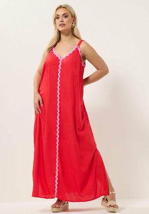 Frau in einem langen roten ärmellosen Kleid mit rosa welligem Besatz, trägt goldene Plateausandalen und steht vor einem schlichten hellen Hintergrund.