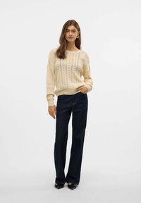 Maglione crema a maglia con motivo a traforato, polsini e orlo a coste, abbinato a jeans svasati blu scuro e décolleté nere a punta.