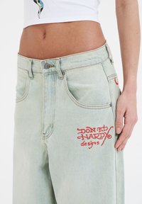 Lichtgewassen denim jeans met een losse fit, voorzien van een rode geborduurde logo aan de voorkant, zilveren knoopsluiting en standaard vijf-pocket ontwerp.