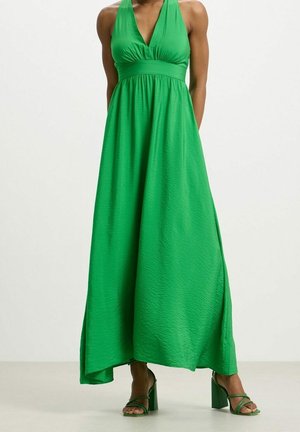 Femme portant une robe maxi sans manches vert vif avec un décolleté en V et des sandales à talons vert assorties, debout sur un fond neutre.
