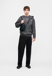 Γκρι φούτερ με φερμουάρ και πλαϊνές τσέπες, φορεμένο πάνω από ένα ασορτί γκρι μπλουζάκι με στρογγυλή λαιμόκοψη. Μαύρο παντελόνι με φαρδιά πόδια και μικρή πράσινη τσάντα χιαστί.