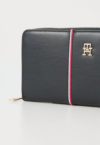 Black leather wallet with a gold logo, featuring a red and white striped accent. Zipper closure with a flat gold hardware detail.