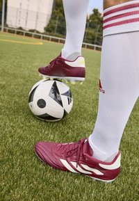 Scarpe da calcio color bordeaux con accenti bianchi e dettagli gialli, posizionate su una palla da calcio con motivo in bianco e nero su erba artificiale.