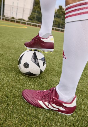 Zapatillas de fútbol color burdeos con acentos blancos y detalles amarillos, colocadas sobre un balón de fútbol con un patrón en blanco y negro sobre hierba artificial.
