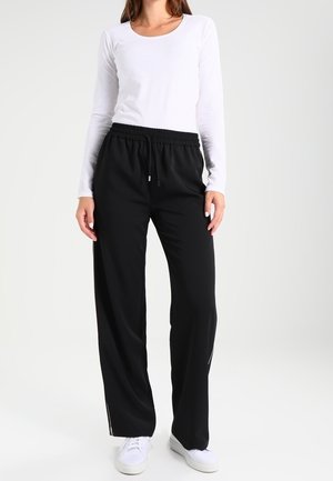Black loose-fitting pants with an elastic waistband and side zipper details, paired with a white long-sleeve top and white sneakers.