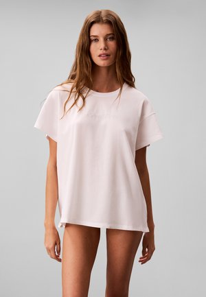 Jonge vrouw met lang bruin haar die een oversized effen lichtroze Calvin Klein T-shirt draagt tegen een effen grijze achtergrond.