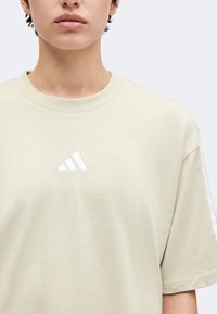 Άτομο φοράει μπεζ μπλούζα Adidas με λευκό λογότυπο στο κέντρο του στήθους, πάνω σε απλό φωτεινό φόντο.