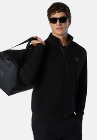 Felpa nera con zip e colletto alto, realizzata in morbido materiale. Presenta tasche laterali e un logo discreto sul petto. Borsa scura appoggiata sulla spalla.