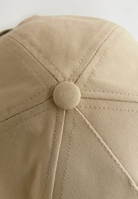 Beige bomull baseballkeps med en strukturerad design, som har en rund krön, sydda paneler och en tygklädd knapp på toppen.