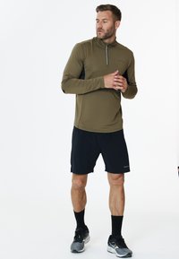 Olivenfarbene Langarm-Sportoberteil mit halbem Reißverschluss, schwarzen Akzenten, kombiniert mit schwarzen Shorts und grauen Sportschuhen, auf einem weißen Hintergrund stehend.