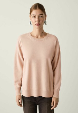 Femme aux cheveux châtains attachés porte un pull rose clair, un pantalon noir et de grandes boucles d'oreilles noires et blanches, debout devant un fond uni.