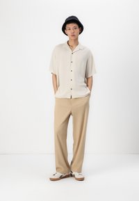 Beige short-sleeve shirt with dark buttons, beige pants, and white sneakers with black accents. The model wears a black bucket hat.